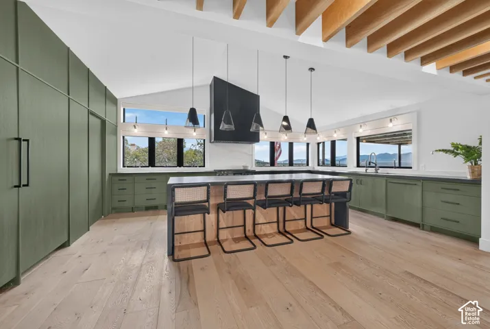 Vaulted ceiling with exposed wood beams, wood-finish flooring, full-height green cabinetry, central island with seating, and multiple windows