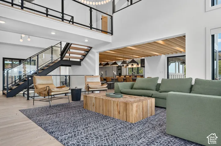 Spacious living area featuring a two-story ceiling, a modern open-tread staircase with glass and metal railings, wood-finish flooring, and exposed wood beam detailing