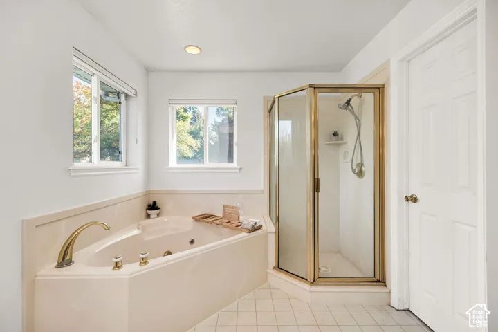 Built-in soaking tub with brass fixtures, individual shower enclosure with glass panels, and light-toned tile flooring