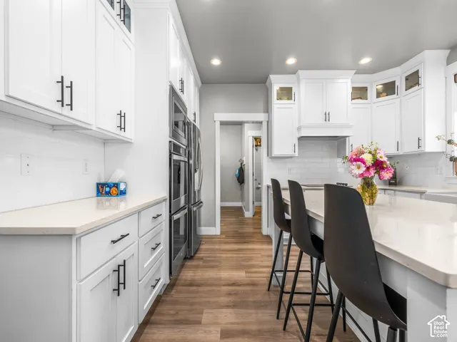 White cabinetry with matte black hardware, light-toned countertops, subway tile backsplash, stainless steel double ovens, and wood-finish flooring