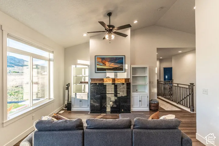 Vaulted ceiling with a ceiling fan, fireplace with a dark stone surround and wood mantle, built-in shelving, large windows, and wood-finish flooring