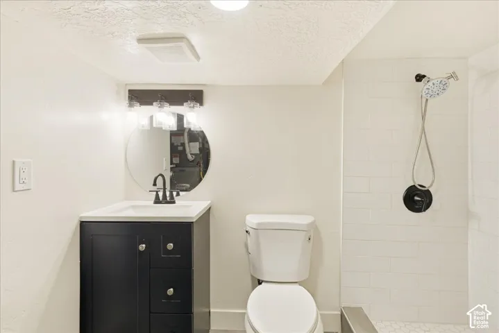 Vanity with dark cabinetry and white countertop, round wall-mounted mirror, three-light fixture with clear glass shades, toilet, and subway-tiled shower with a handheld showerhead