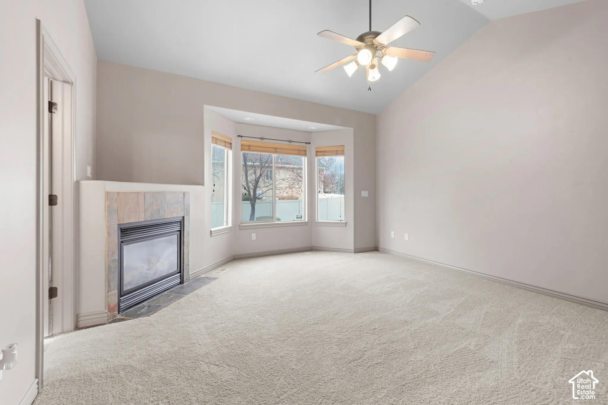 Vaulted ceiling with ceiling fan, bay window with blinds, carpet flooring, and a fireplace with a stone surround