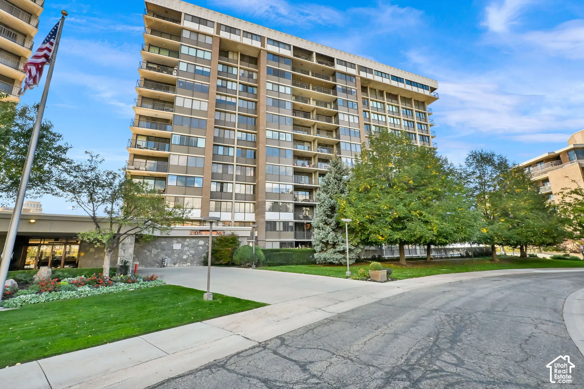 Modern mid-rise residential building featuring multiple levels of unit windows, a landscaped grounds with mature trees and manicured lawn, concrete walkways, and an American flag at the entrance