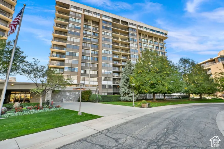 Modern mid-rise residential building featuring multiple levels of unit windows, a landscaped grounds with mature trees and manicured lawn, concrete walkways, and an American flag at the entrance
