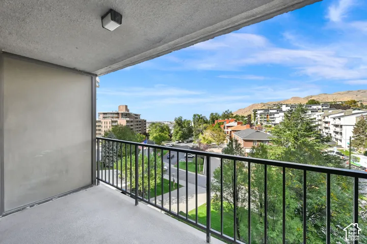 Balcony with metal railing and textured flooring, featuring a wall-mounted light fixture and surrounding cityscape