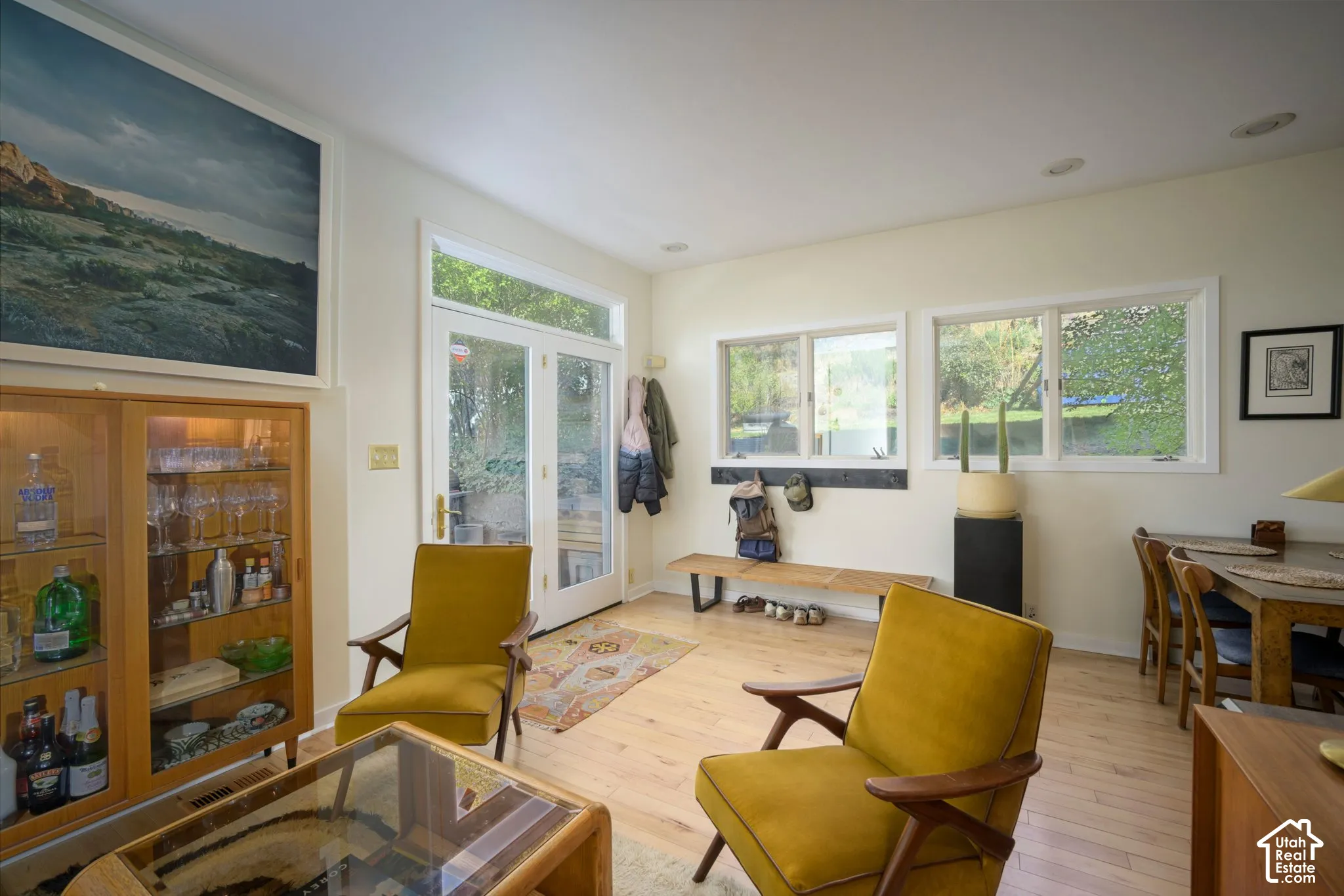 Light wood-finish flooring throughout, double glass-paneled doors with transom, multiple windows providing ample ambient light, recessed ceiling lighting, and a built-in display cabinet