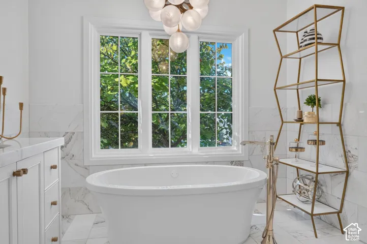 Freestanding soaking tub with floor-mounted faucet, white shaker cabinetry with brass hardware, and wall-to-wall stone-look tile