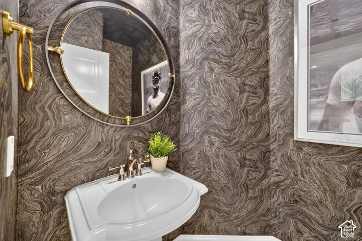 Pedestal sink with polished nickel faucet, round mirror with brass accents, and patterned wall covering