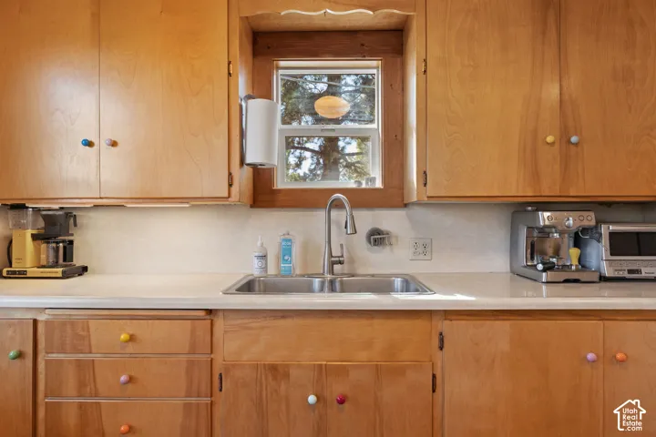 Wood-finish cabinetry with colorful knobs, dual basin stainless steel sink with a gooseneck faucet, light-toned countertop, and a window with wood trim