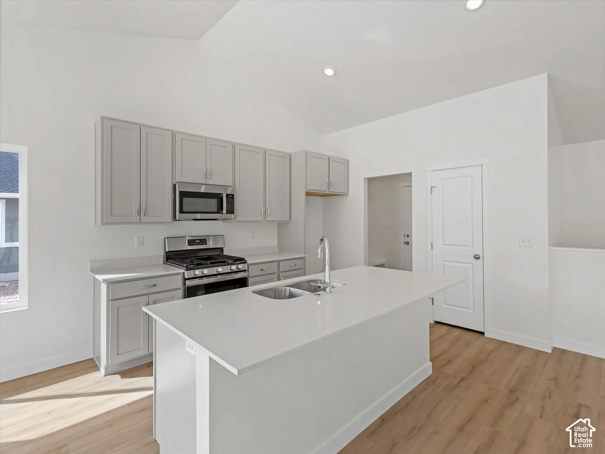Kitchen featuring vaulted ceilings, light wood-finish flooring, light gray cabinetry, stainless steel appliances, and a central island with an undermount sink