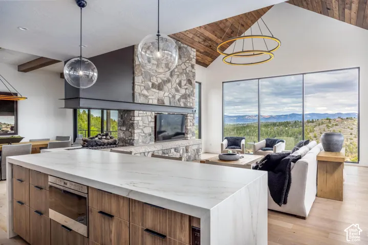Vaulted ceilings with wood-finish accents, expansive floor-to-ceiling windows, a stone fireplace, a large kitchen island with a built-in appliance, and modern pendant lighting
