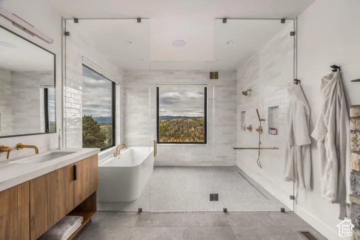 Dual vanity with wood-finish cabinetry, a freestanding soaking tub, a glass-enclosed shower, expansive windows, and light-toned tile flooring