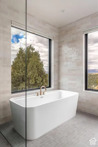 Freestanding soaking tub with bronze-finish fixtures, large black-framed windows, light-toned wall tile, and gray floor tile