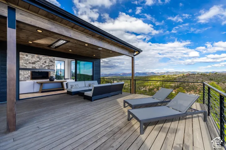Expansive outdoor deck with wood-finish flooring, cable railing, and a covered patio featuring a stone-clad fireplace and recessed lighting