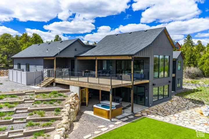 Contemporary exterior with board and batten siding, expansive windows, a large wood-finish deck with cable railing, and a ground-level patio featuring a hot tub