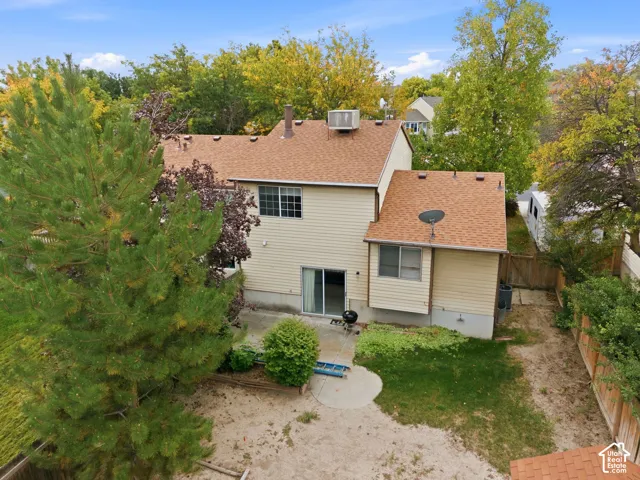 Rear exterior featuring a sliding glass door, a concrete patio, a shingled roof, mature trees, and a fenced perimeter