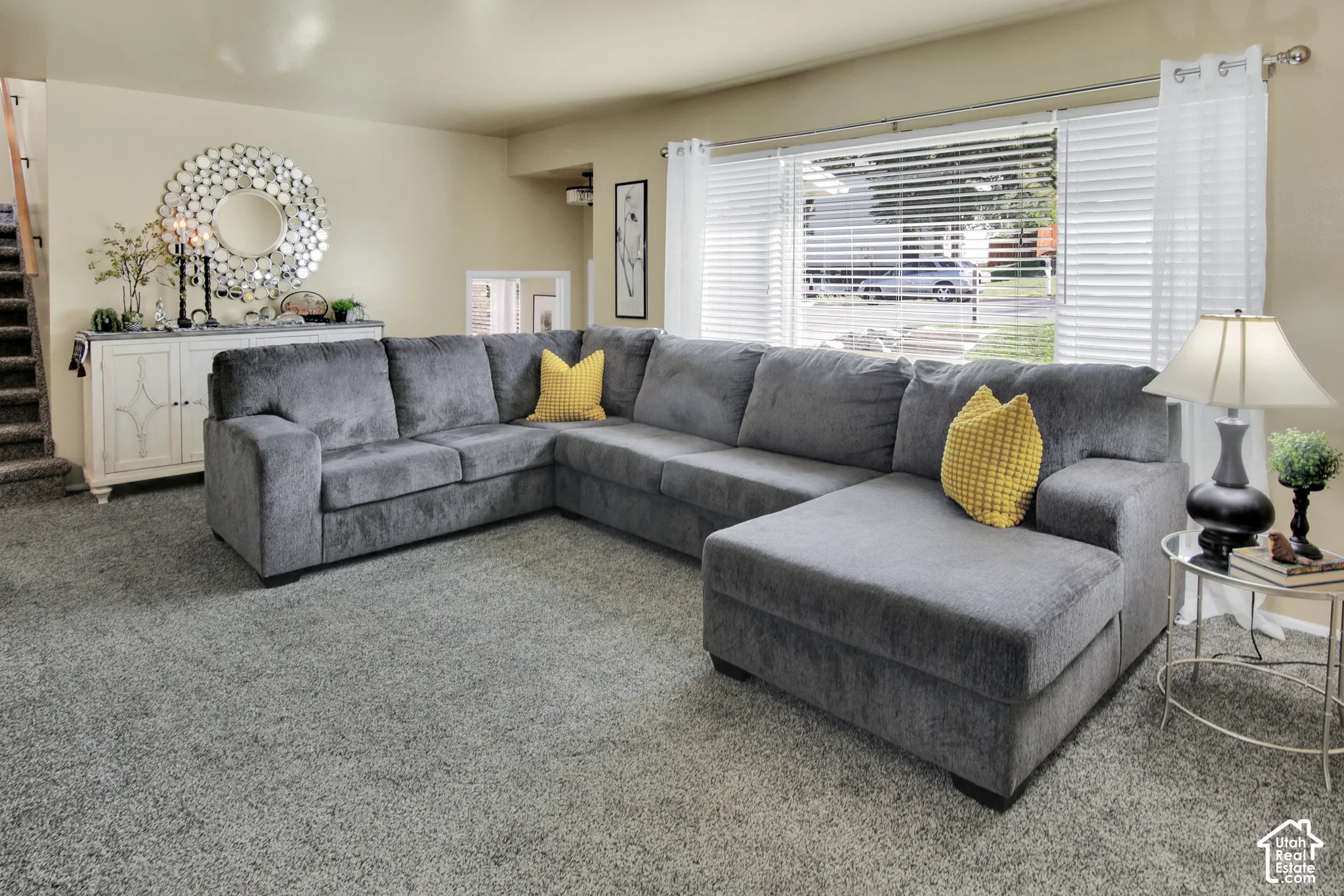 Living area featuring soft neutral carpeting, a large window with horizontal blinds, and a built-in stairwell