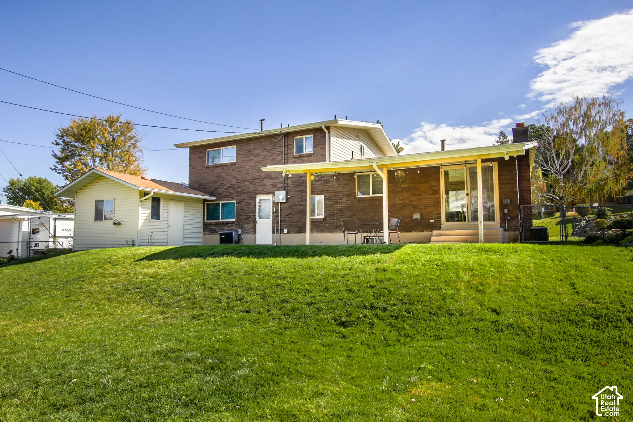 Expansive grass-covered lawn with a two-story brick exterior, attached covered patio, storage shed, and mature landscaping