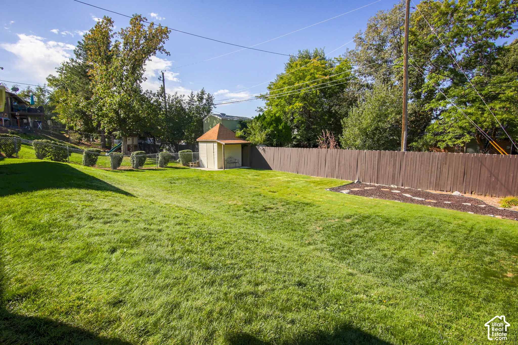 Expansive lawn area with a privacy fence, mature trees, manicured shrubs, and a storage shed