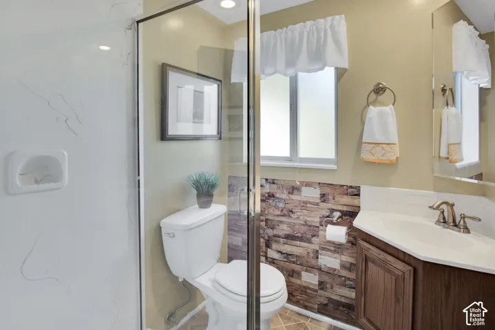 Glass-enclosed shower with white-patterned wall, corner vanity with wood-finish cabinetry, white countertop, and single-handle faucet, and a toilet with an adjacent stone-patterned wall