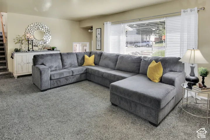Living area featuring soft neutral carpeting, a large window with horizontal blinds, and a built-in stairwell