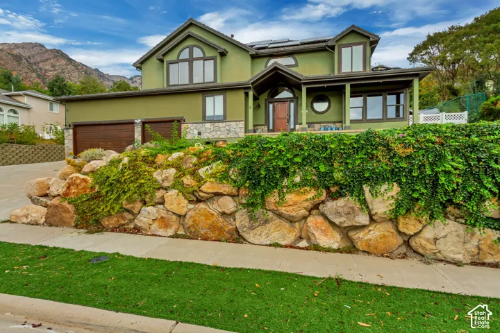 Stucco exterior featuring stone accents, wood-finish garage doors, and a decorative rock retaining wall with lush greenery