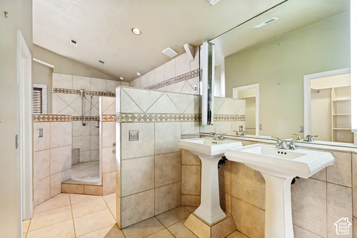 Dual pedestal sinks, full wall-length mirror, tiled flooring, tiled walk-in shower with built-in bench, and vaulted ceiling with recessed lighting