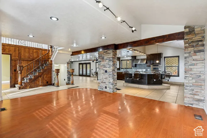 Wood-finish flooring, stone-clad columns, a grand wooden staircase, recessed ceiling lighting, and a kitchen with a large island