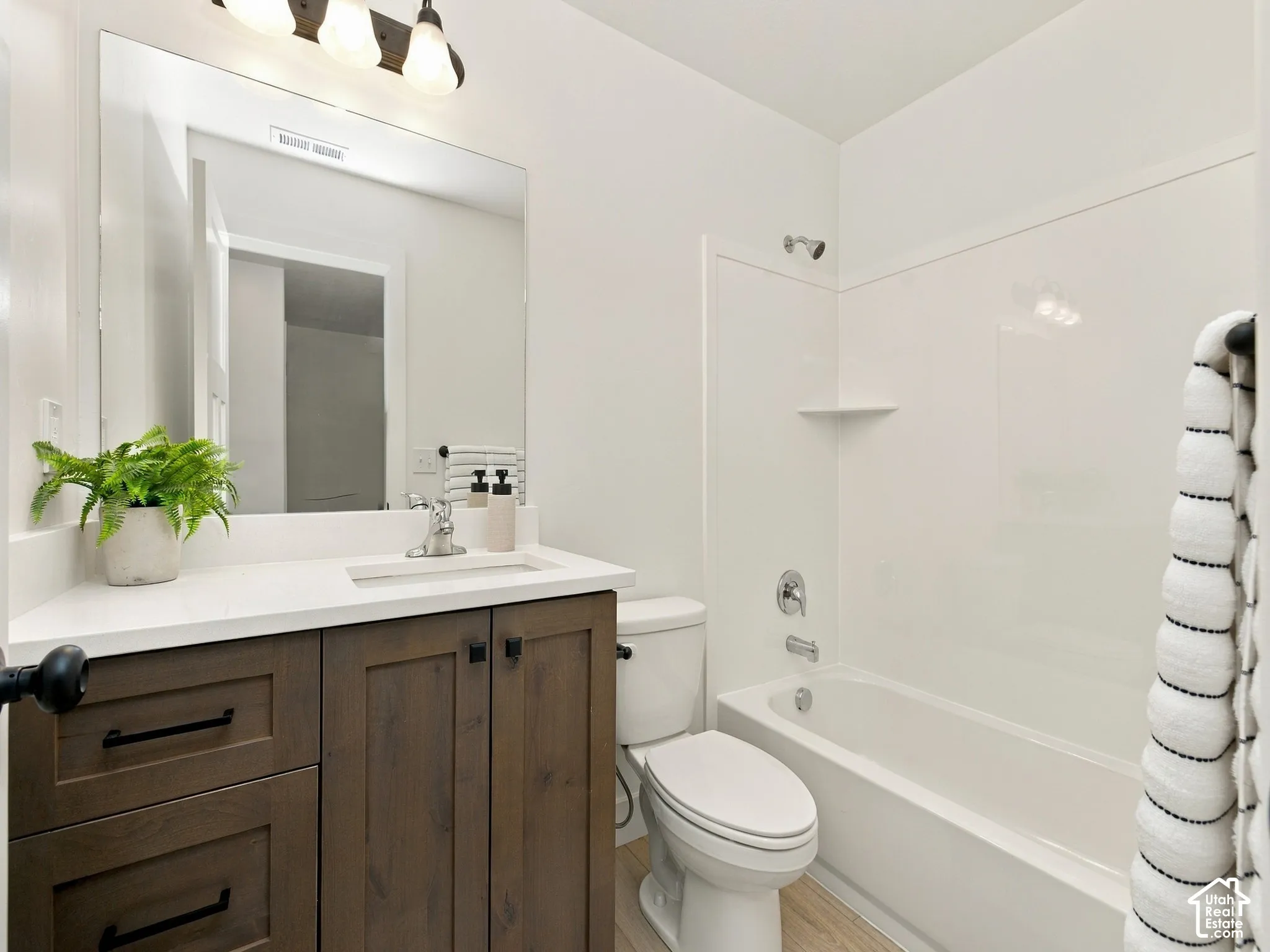 Bathroom featuring a wood-finish vanity with dark hardware, a white countertop with an integrated sink, a rectangular mirror, a three-light fixture, and a bathtub with a shower