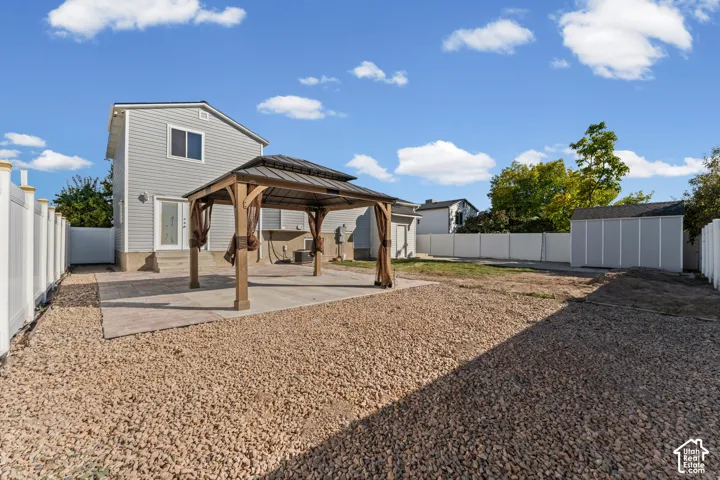 Expansive backyard featuring a gravel landscape, a concrete patio, a wooden gazebo with a metal roof, a white vinyl privacy fence, and an exterior storage shed