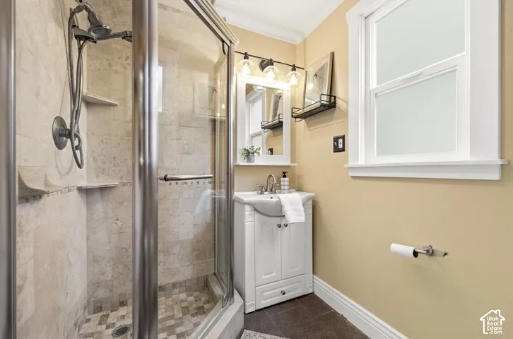 Glass-enclosed shower with stone tile surround and built-in shelving, white vanity with integrated sink, window with white trim, and a three-light vanity fixture