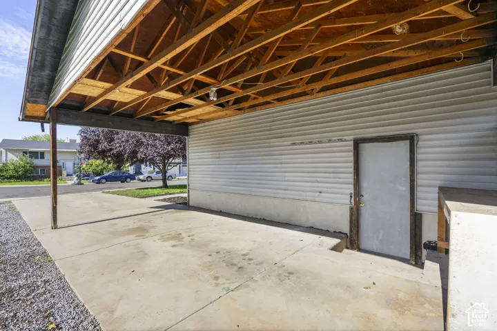 Covered concrete parking area with exposed wood ceiling joists and a side entry door