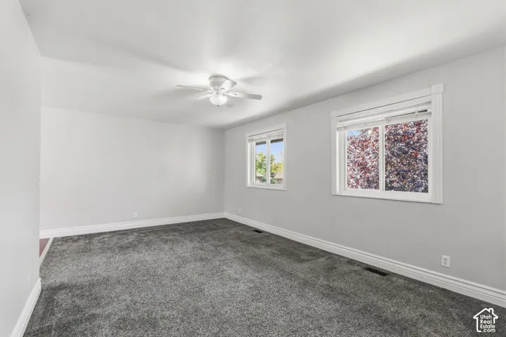 Light gray walls with white trim, gray carpeted flooring, two windows, and a ceiling fan