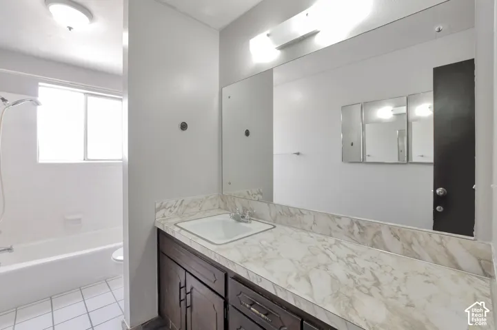 Vanity with a faux marble countertop and an integrated sink, a large wall-mounted mirror, dark wood-finish cabinetry with silver-tone hardware, a shower-tub combination, and tiled flooring