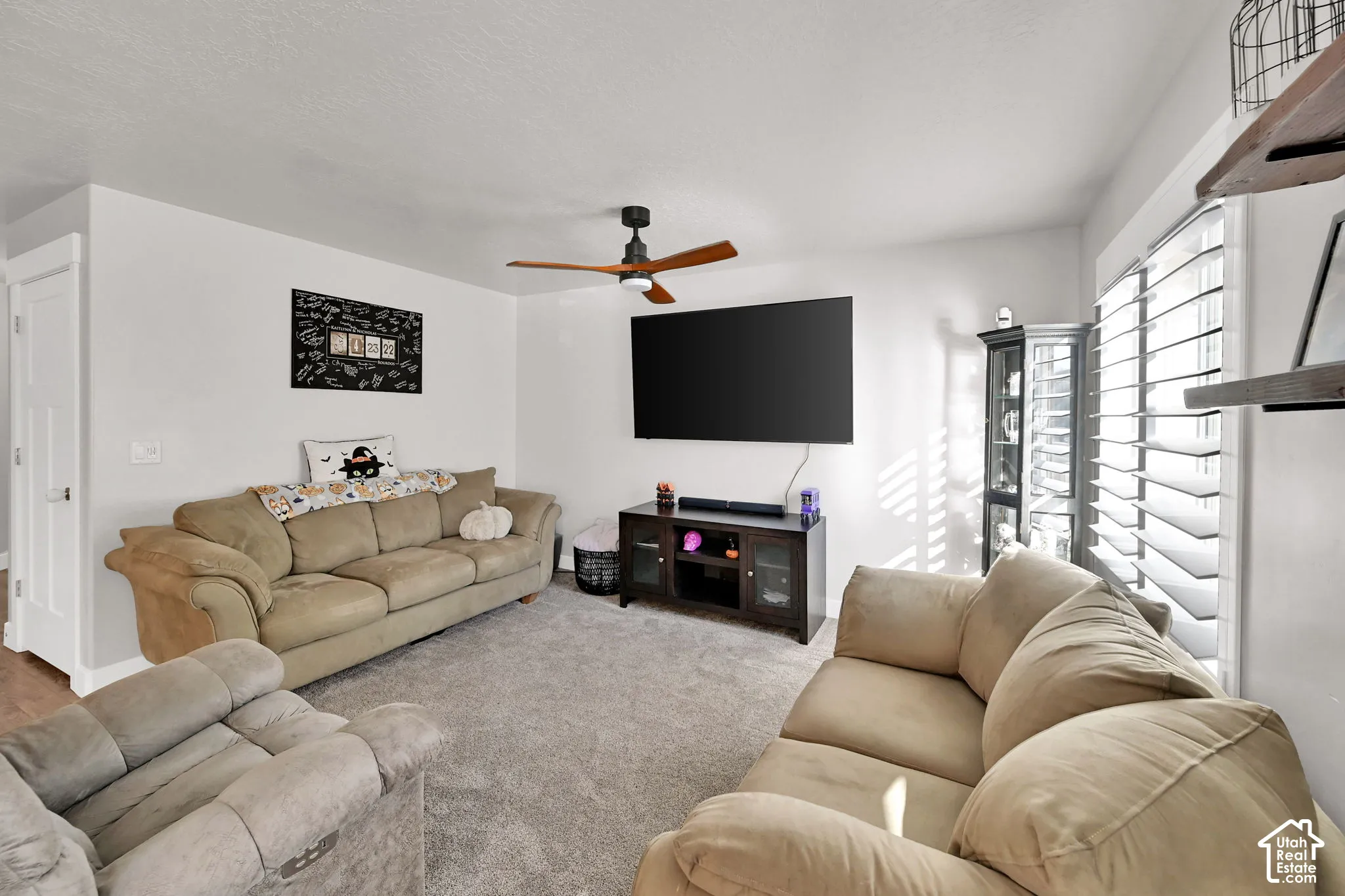 Carpeted living area featuring a ceiling fan, white walls, and a window with white louvered shutters