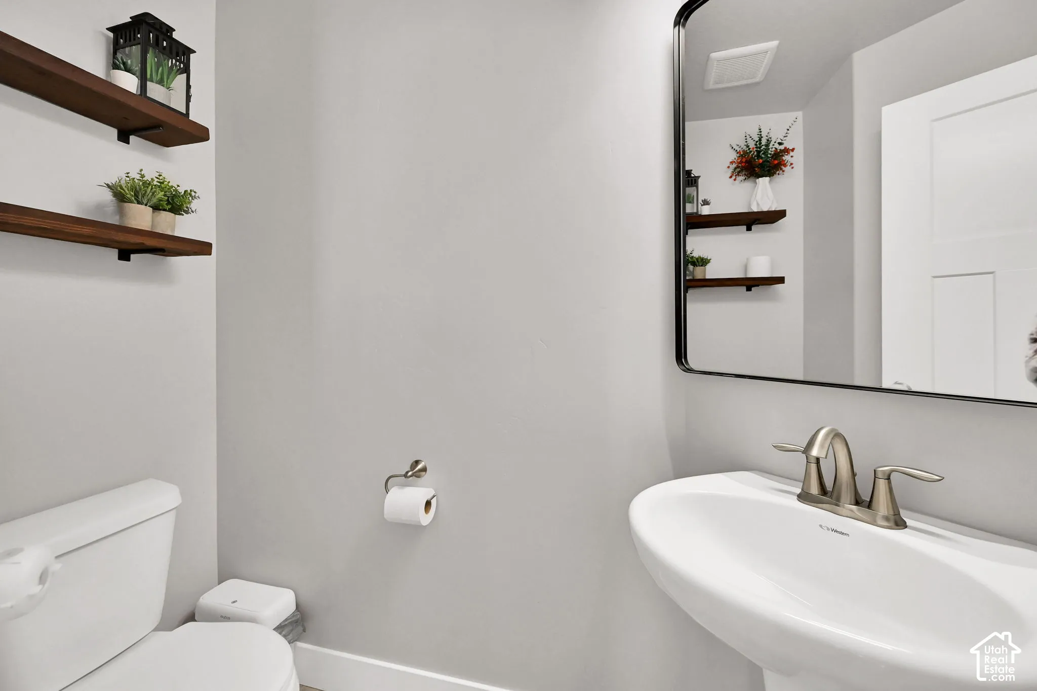 Pedestal sink with brushed metal faucet, rectangular framed mirror, and two wall-mounted wood-finish shelves