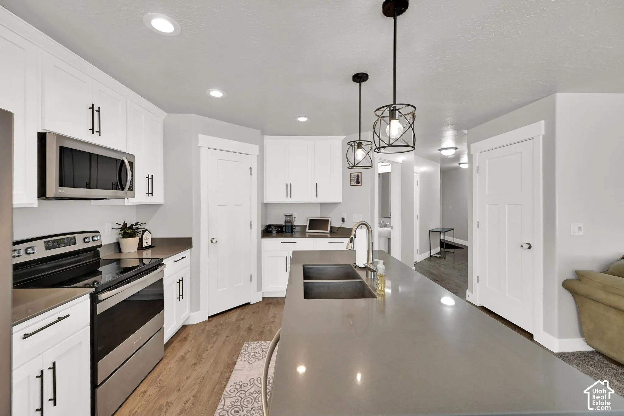 White cabinetry with matte black hardware, stainless steel appliances, wood-finish flooring, recessed lighting, and an island with a double basin sink