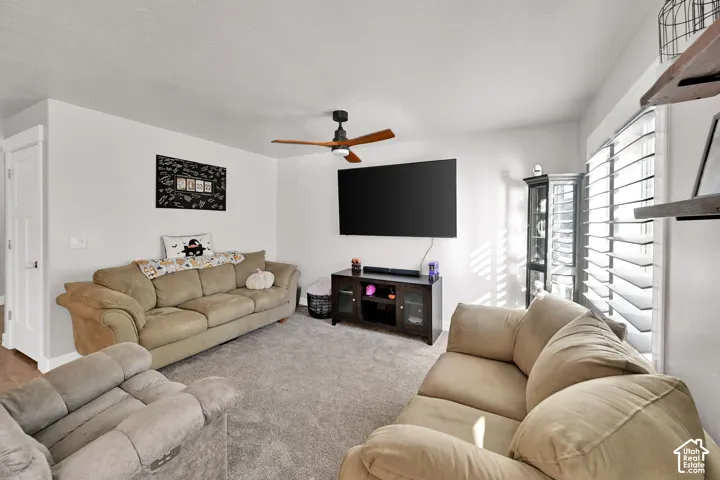 Carpeted living area featuring a ceiling fan, white walls, and a window with white louvered shutters