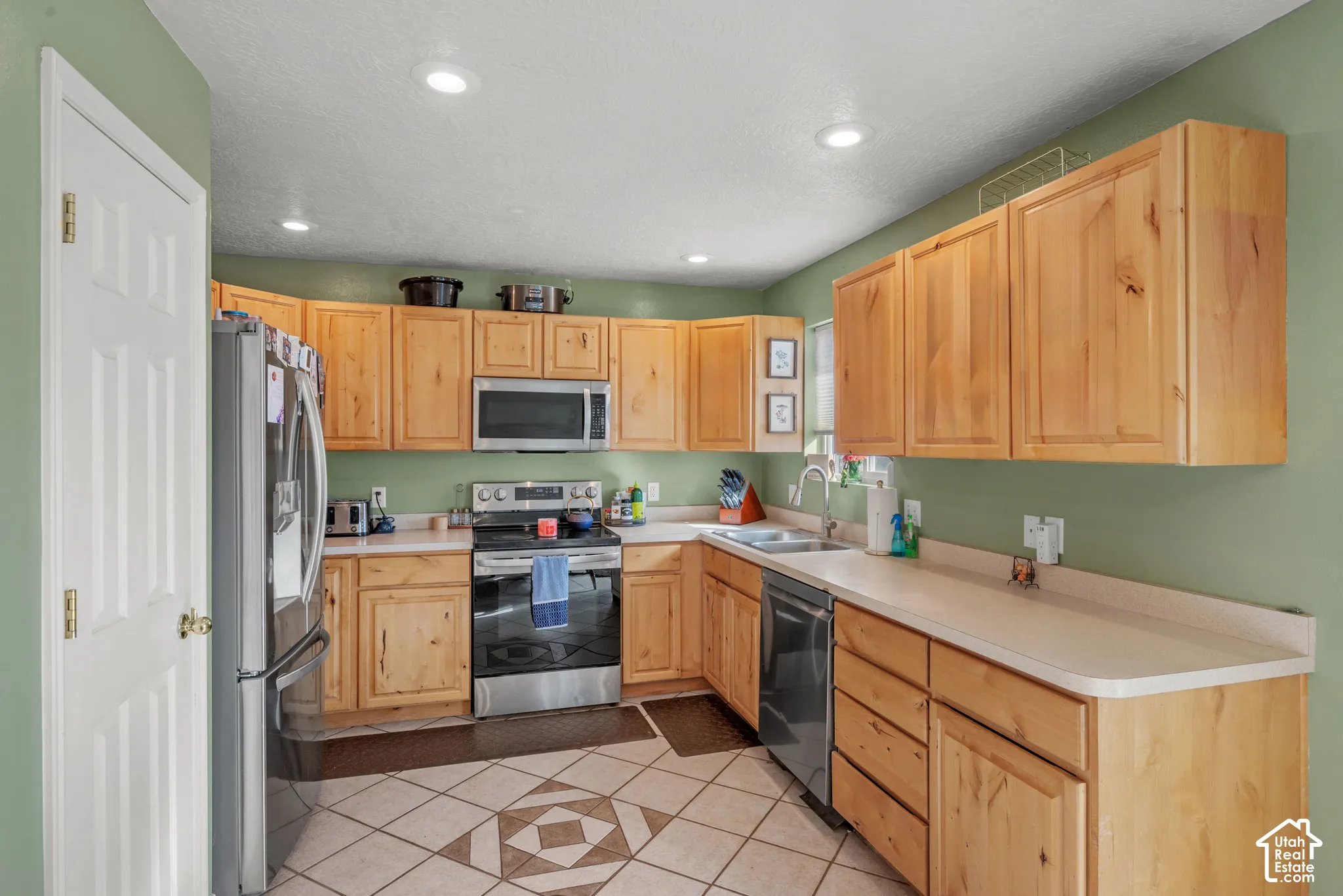 Well-appointed kitchen featuring light wood cabinetry, light-toned countertops, tile flooring with geometric patterns, recessed lighting, and stainless steel appliances