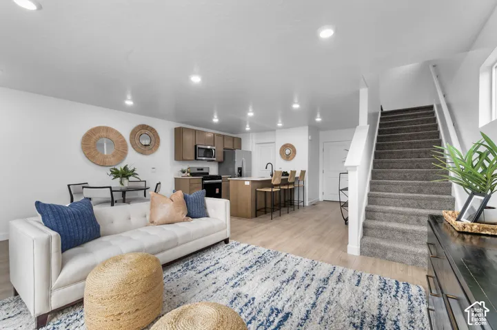 Open-concept living area featuring wood-finish flooring, recessed lighting, a modern kitchen with wood cabinetry and stainless steel appliances, and a carpeted staircase with white railings