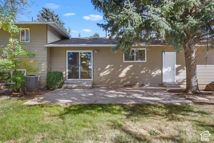 Rear exterior featuring a concrete patio, sliding glass door, tan horizontal siding, and a mature tree