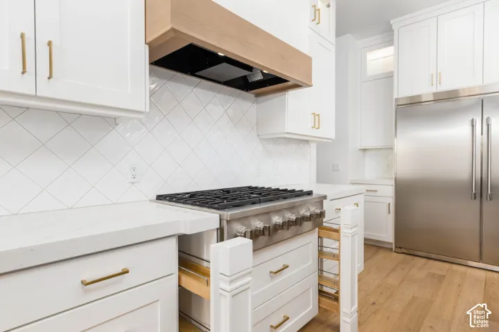 Professional-grade stainless steel range with gas burners, white shaker cabinetry, custom wood-finish range hood, white tile backsplash, and stainless steel refrigerator