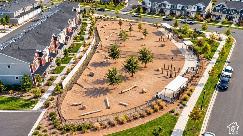 Community park featuring a generous sand play area with integrated playground equipment, mature shade trees strategically planted throughout, curved hardscape pathways, and perimeter landscaping with ornamental plantings