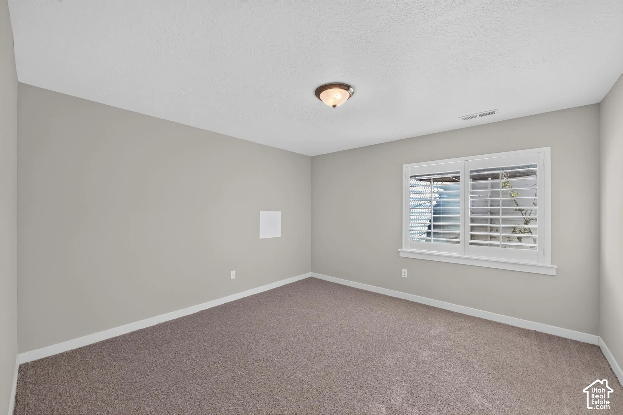 Neutral wall paint, textured carpeting, window with plantation shutters, white trim, and a ceiling-mounted light fixture