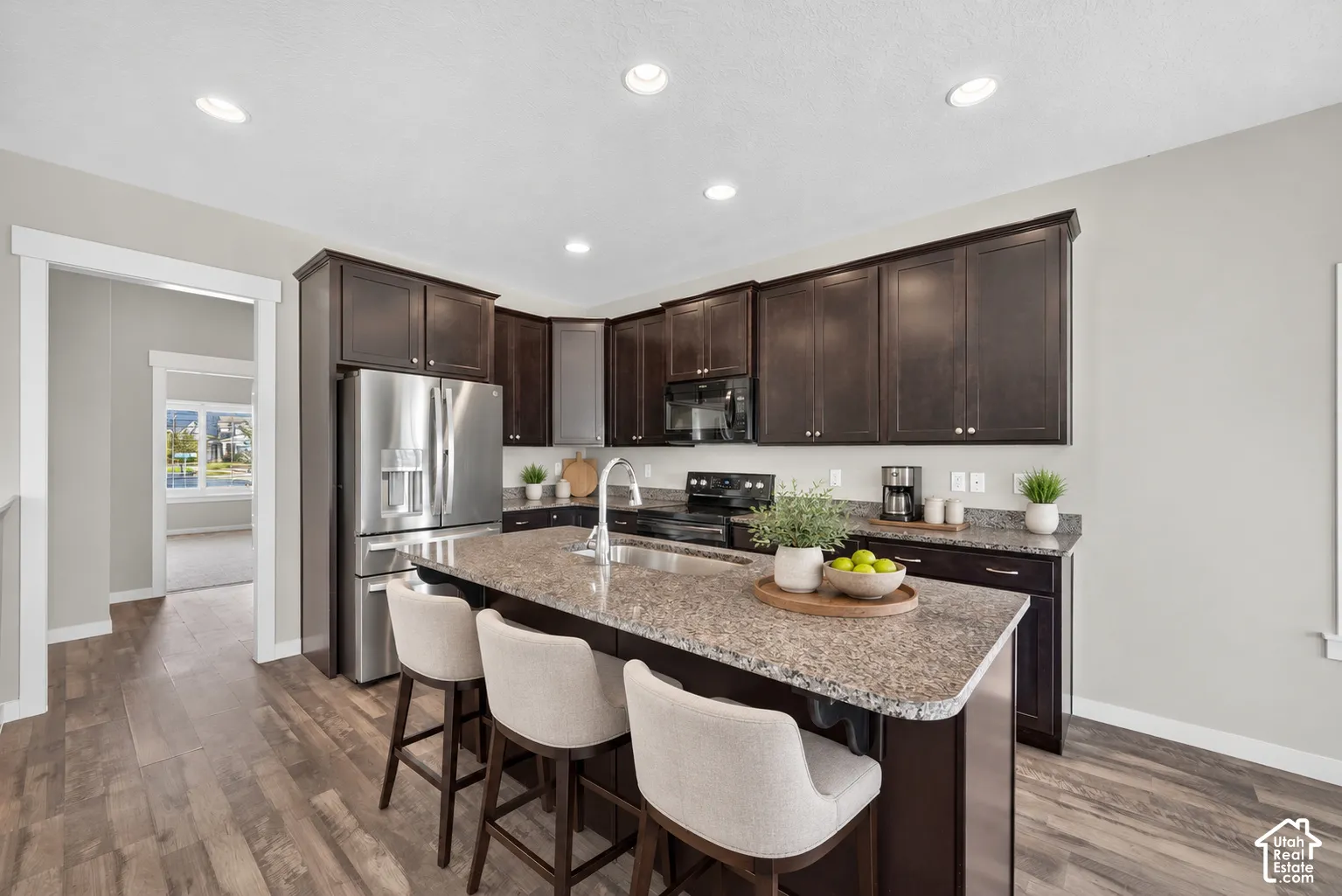 Modern kitchen featuring dark cabinetry with ample storage, stainless steel appliances including a refrigerator and range, granite countertops, a large island with seating for multiple guests, recessed ceiling lighting, and wood-finish flooring throughout