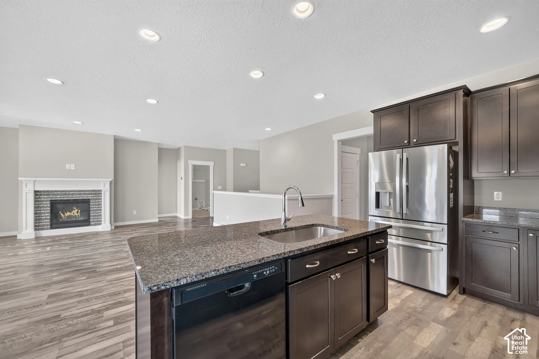 Kitchen island with stone countertop and integrated sink, stainless steel refrigerator, dark wood-finish cabinetry, wood-finish flooring, and a fireplace with a tile surround