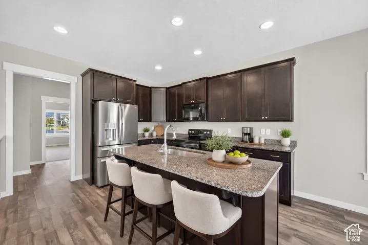 Modern kitchen featuring dark cabinetry with ample storage, stainless steel appliances including a refrigerator and range, granite countertops, a large island with seating for multiple guests, recessed ceiling lighting, and wood-finish flooring throughout