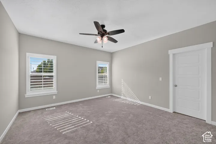 Neutral tone carpeted flooring, two windows with white plantation shutters, a ceiling fan with integrated lighting, and a white paneled door