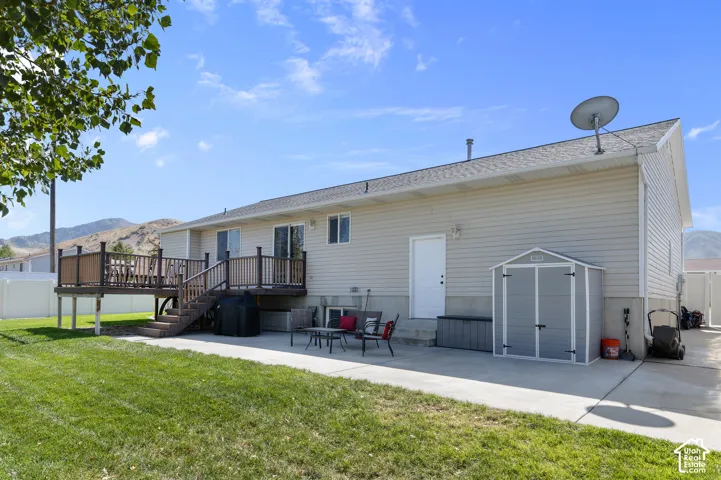 Spacious manufactured home featuring a composite deck with integrated seating areas, expansive manicured lawn with mature shade trees, detached storage shed, concrete pad foundation, and mountain backdrop